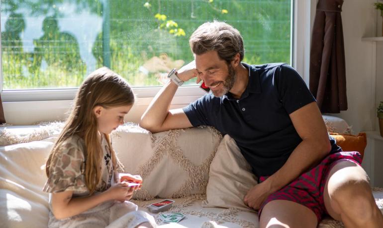 Bambina e uomo giocano a carte su un divano soleggiato.